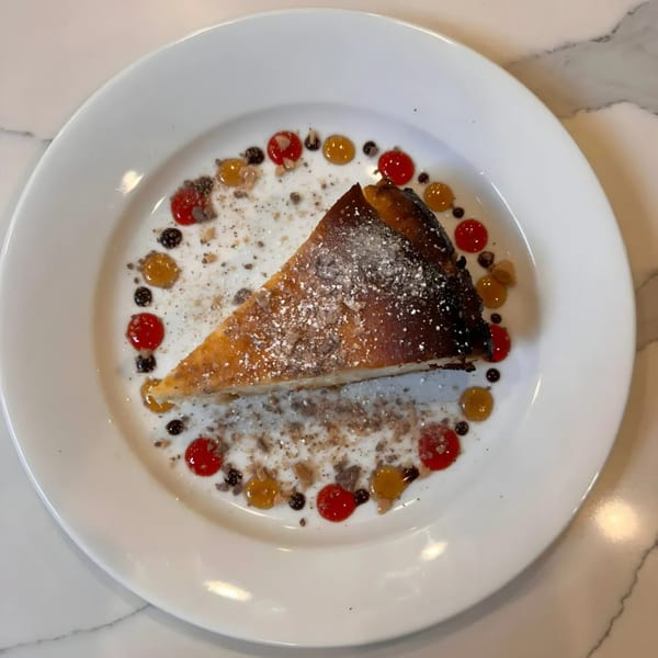 A slice of cheesecake with powdered sugar, surrounded by colorful fruit purees and crumbles on a white plate.